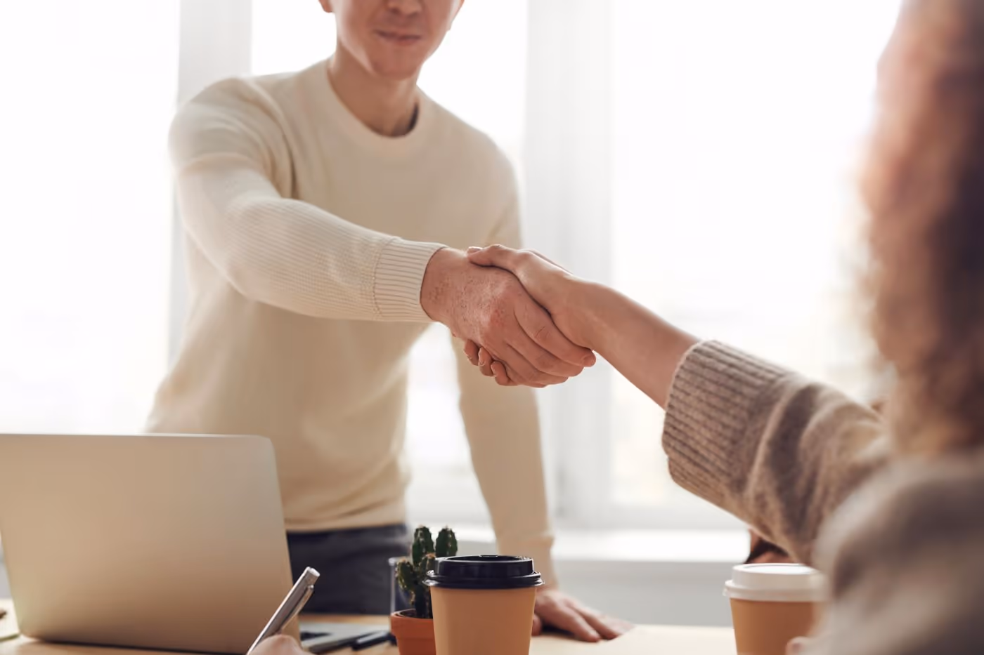 Two people shaking hands across a desk with a laptop