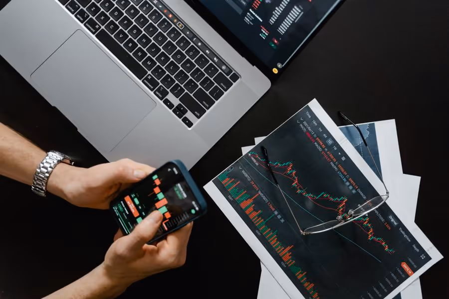 Hands holding a phone with a trading chart next to a laptop and printed charts